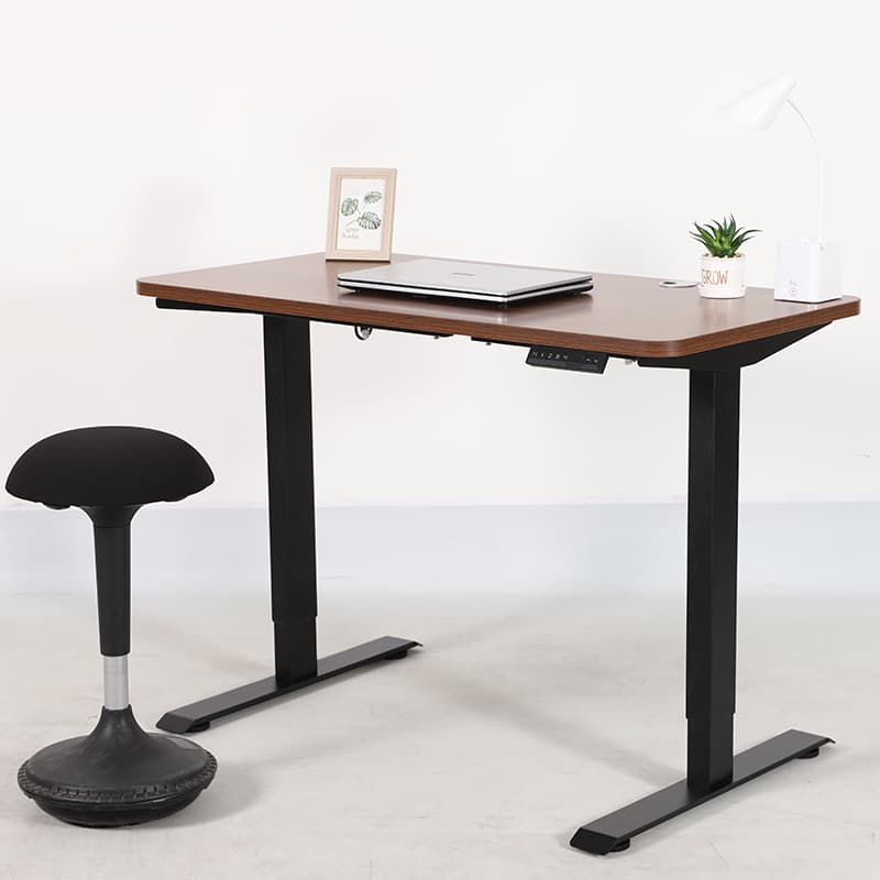 A fully assembled 2AR2 adjustable standing desk with a black frame and a dark wood tabletop, shown in a room setting with a laptop, lamp, and a wobble stool.