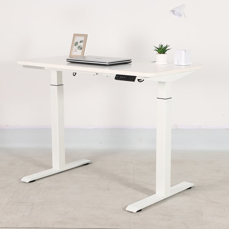 White 2B2 adjustable standing desk with laptop, plant, and lamp on a light gray floor against a white wall.